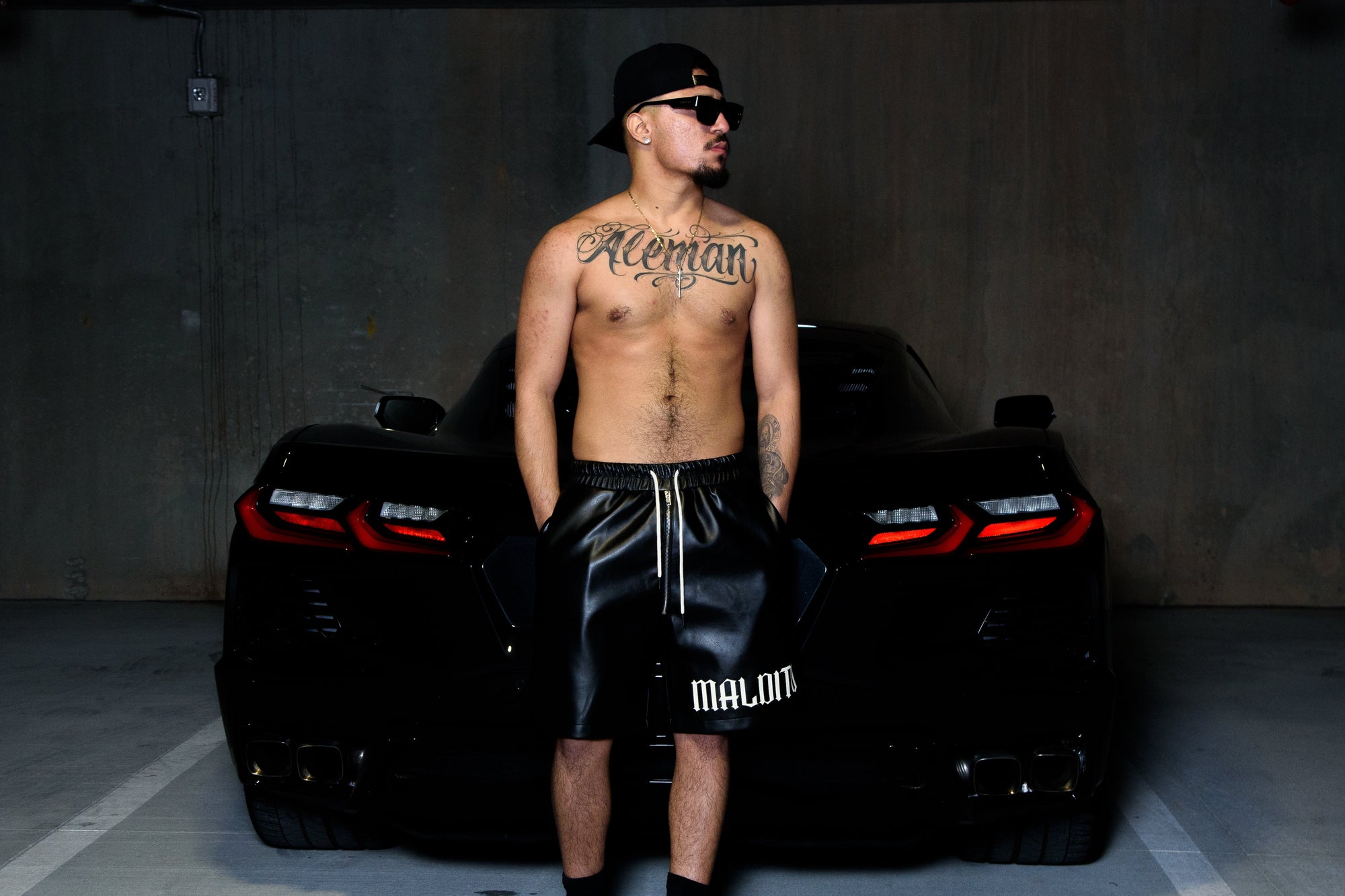 Shirtless model wearing MALDITO leather shorts standing in front of a sports car for the brand’s hero campaign image